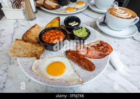 Traditionelles englisches Frühstück serviert auf einem Marmortisch mit gebratenem Ei, Speck, Wurst, gebackenen Bohnen, Toast, Avocado und Kaffee in einem Café. Stockfoto