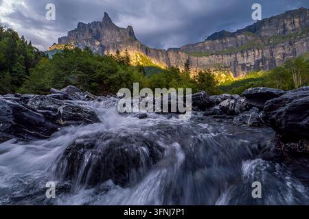 Ein stürmender Gebirgsbach fließt über dunkle Felsen unter den dramatischen Aiguilles Rouges-Klippen in den französischen Alpen, wo warmes Sonnenlicht durchbricht Stockfoto