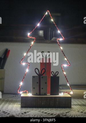 Eine Nahaufnahme eines DIY-Weihnachtsbaums aus Draht und leuchtenden Lichtern im Innenraum, begleitet von drei kleinen handgefertigten Holzgeschenkboxen auf einem strukturierten Ta Stockfoto