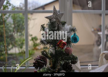 Detaillierte Aufnahme der Spitze eines kleinen künstlichen Weihnachtsbaums, der mit einem silbernen Stern, einer glänzenden blauen Kugel, einer Miniaturgeschenkbox und schneeartigem Cotto verziert ist Stockfoto