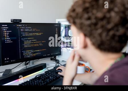 Rückansicht eines jungen Mannes, der auf einem Desktop-Monitor an einem Gaming-Computer-Setup Code schreibt und dabei KI-Werkzeuge für die Softwareentwicklung in einem modernen Heimarbeitsplatz verwendet Stockfoto