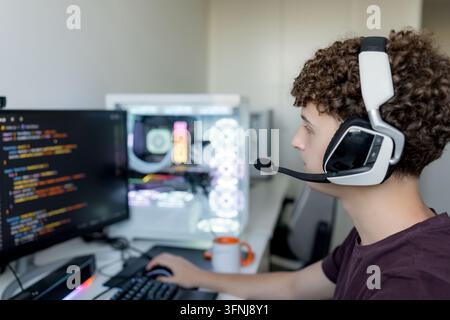 Junger Mann mit Headset nutzt KI-Tools und einen Gaming-Computer zu Hause, betrachtet Code auf einem Monitor neben einem beleuchteten Desktop-PC. Stockfoto