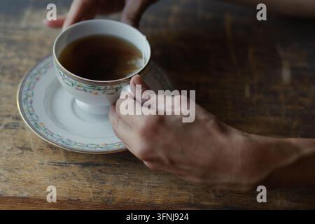Menschliche Hände halten eine Vintage-Porzellan-Teetasse, gefüllt mit heißem schwarzem Tee, auf einem dunklen rustikalen Holztisch Stockfoto