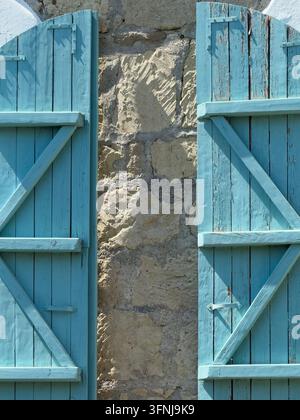Nahaufnahme von verwitterten blauen Holzläden, die eine raue Steinfaçade einrahmen, mit alternder Farbe, stabilen Streben, Metallscharnieren und traditioneller Architektur Stockfoto