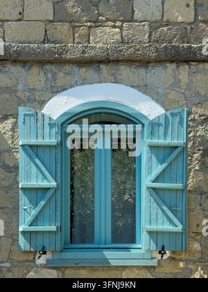 Helle türkisfarbene Holzläden rahmen ein gewölbtes Fenster, das in eine verwitterte Steinfassade eingelassen ist, und schaffen so ein bezauberndes architektonisches Detail im Sonnenlicht. Stockfoto