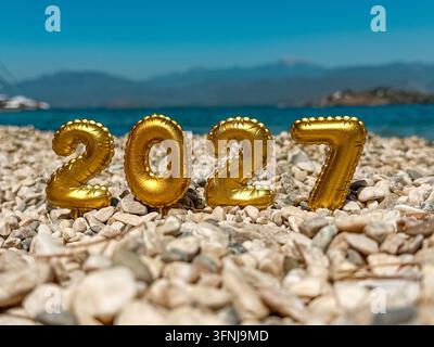 Glänzende Goldfolienballons, die die Zahl 2027 bilden, stehen an einem Kieselstrand neben dem Meer in der Dämmerung, mit weichem blauem Himmel und Wellen, die eine festliche Neujahrsfeier schaffen Stockfoto