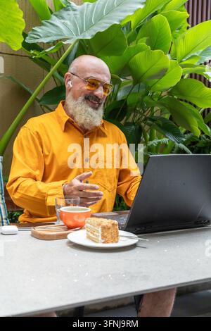Lächelnder Senior-Unternehmer in einem orangefarbenen Hemd verwendet einen Laptop und einen kabellosen Ohrhörer an einem Gartentisch, genießt Kaffee und Kuchen, während er aus der Ferne arbeitet Stockfoto