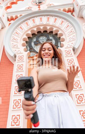 Junge Frau nimmt sich selbst mit einer Kompaktkamera auf, während sie vor einer dekorativen gewölbten Fassade steht, und erstellt Reiseinhalte für einen Vlog, Social Media Stockfoto