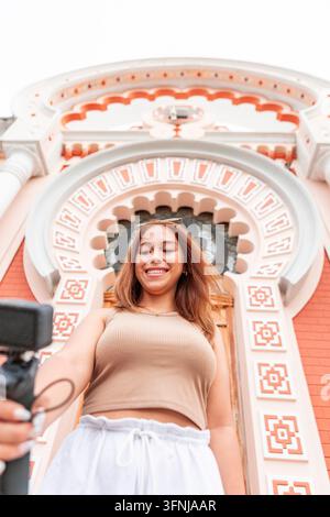 Lächelnde junge Frau filmt sich selbst mit einer Handkamera vor einer verzierten historischen Fassade und erstellt Reiseinhalte für einen Vlog, soziale Medien oder um Stockfoto