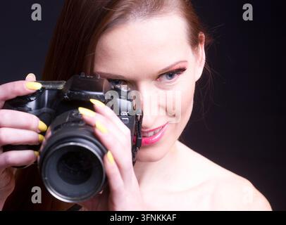 Eine Fotografin zielt mit einer DSLR-Kamera in einem Studio-Porträt und zeigt Selbstvertrauen, Kreativität und professionelle Fotografie mit hellgelben Nägeln Stockfoto
