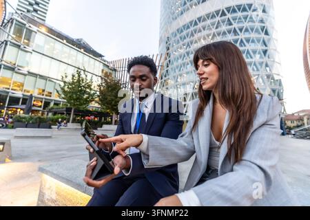 Zwei vielfältige Geschäftsleute arbeiten gemeinsam im Freien an einem digitalen Tablet zwischen modernen Wolkenkratzern, teilen Ideen, planen Strategien und nutzen Technologie Stockfoto
