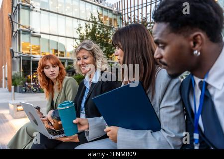 Verschiedene Wirtschaftsexperten treffen sich lässig im Freien, arbeiten gemeinsam an einem Laptop und teilen Ideen, was Teamarbeit und moderne Arbeitskultur repräsentiert Stockfoto