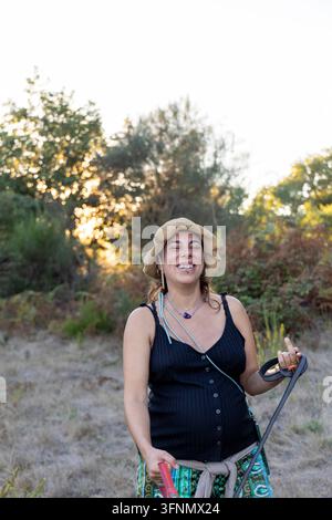 Schwangere hispanische Frau mit einem freudigen Ausdruck, trägt einen Hut und ein schwarzes Oberteil, steht in der Natur bei Sonnenuntergang Stockfoto