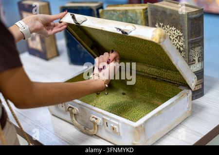 Frau öffnet einen Vintage-Koffer auf einem Tisch, umgeben von dekorativen Büchern, antiker Reisekoffer mit Retro-Interieur und nostalgischem Design. Stockfoto