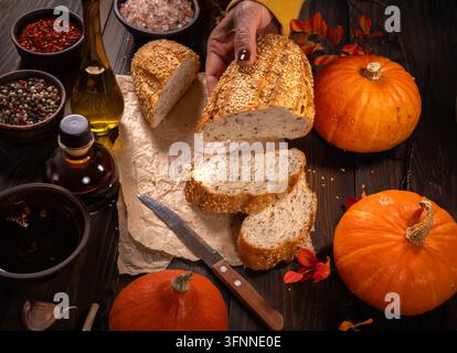 Hand, die frisch gebackenes Brot mit Samen hält, auf einem rustikalen Holztisch geschnitten, mit ganzen Kürbissen, verschiedenen Gewürzen und Ölflaschen, um ein gesundes Essen zuzubereiten Stockfoto