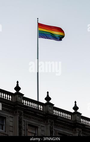Vertikale Niedrigwinkelaufnahme einer lebendigen Regenbogen-Pride-Flagge, die an einem hohen Flaggenmast über einem verzierten klassischen Steinbalkon flattert. Die farbenfrohe Stockfoto