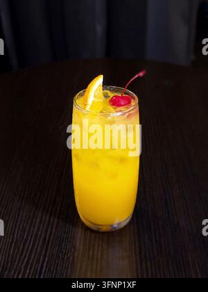 Erfrischender Orangencocktail mit Eis, garniert mit einer Orangenscheibe und einer roten Kirsche, serviert in einem hohen Glas auf einem dunklen Holztisch. Stockfoto