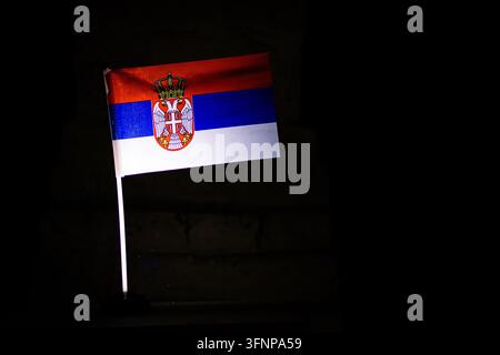Die serbische Trikolore mit Wappen fängt ein starkes Licht auf einem dunklen Hintergrund ein, wobei das Rot, Blau und Weiß sanft weht, um das Erbe der Widerstandsfähigkeit darzustellen Stockfoto
