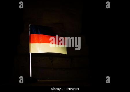 Deutscher Tricolor in Schwarz, Rot und Gold leuchtet vor einem tiefschwarzen Hintergrund, der Stoff weht sanft und reflektiert Einheit, Demokratie, Fleiß und Erbe Stockfoto