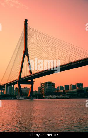 SHANGHAI, CHINA - 16. JUNI 2025: Niedrige Ansicht eines ikonischen A-förmigen Kabelstützenpfeilers und Stahlseile, die sich vor einem lebendigen orangefarbenen Himmel abheben Stockfoto