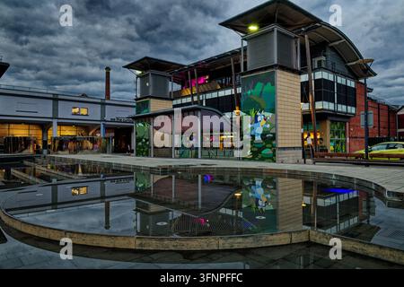 Ein Wasserspiel in einem öffentlichen Platz in der Dämmerung, während sich Abendwolken über Bars und Restaurants mit aufgehenden Lichtern sammeln, Millennium Square, Bristol. Stockfoto