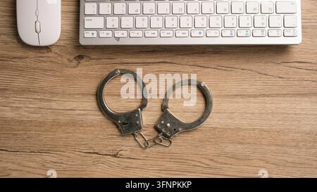 Metallhandschellen auf Holzschreibtisch neben Computertastatur und Maus, die Cyberkriminalität, Cybercrime-Konzepte, Online-Betrug, Internetsicherheit und Digitales repräsentieren Stockfoto