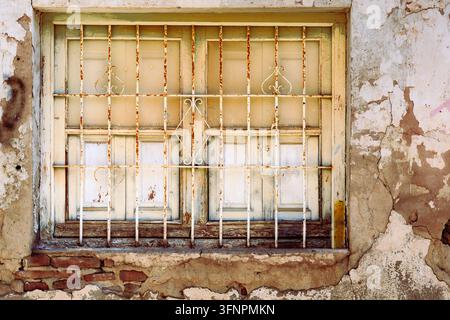 Ein altes Holzfenster mit geschlossenen Fensterläden und abblätternder Farbe ist von rostigen weißen Metall-Sicherheitsgittern bedeckt, die dekorative Schnörkelmuster enthalten Stockfoto
