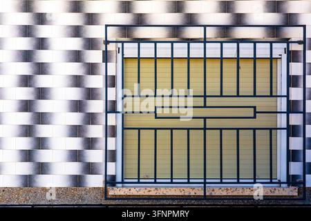 Die Fassade eines Gebäudes zeigt eine Wand, die mit schwarz-weißen, rechteckigen Kacheln in Farbverlauf neben einem Fenster mit geschlossenem gelbem horizontalem Rollos versehen ist Stockfoto