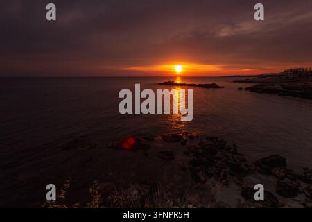 Dramatischer orangefarbener Sonnenuntergang über einem ruhigen Meer mit einem leuchtenden Spiegelbild auf dem Wasser und einer dunklen felsigen Küste unter einem bewölkten Abendhimmel. Stockfoto