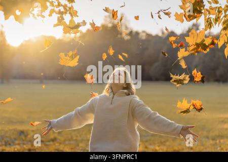 Lächelnde Frau steht mit offenen Armen zwischen vielen fallenden Ahornblättern im warmen Licht der goldenen Stunde. Herbstfreude, Freiheit, unbeschwerte Stimmung, positive Emotionen Stockfoto