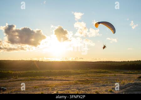 Weite Panoramasicht eines motorisierten Gleitschirmfliegers, der während des Sonnenuntergangs über offene Felder und den fernen Waldhorizont schwebt. Warmes goldenes Sonnenlicht f Stockfoto