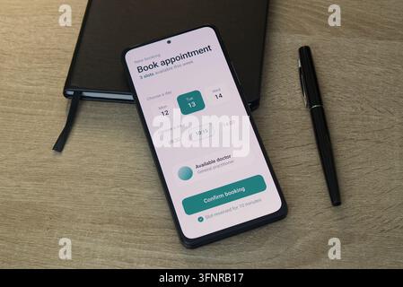 Smartphone, das eine Benutzeroberfläche einer medizinischen Terminbuchungs-App mit Datums- und Uhrzeitauswahl anzeigt, neben einem schwarzen Leder-Notizbuch und einem Stift auf einem Tisch Stockfoto