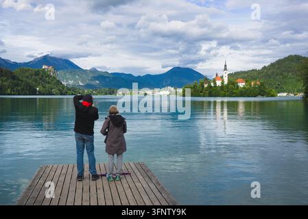 Zwei Touristen stehen auf einem Holzsteg und genießen den malerischen Blick auf den Bledsee, die Bleder Insel und das Bleder Schloss in Slowenien. Stockfoto