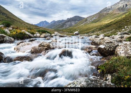 Bergbach stürzt über Felsen im Tal. Das Wasser fließt schnell, weißer Schaum umgibt die Steine. Grüne Wiesen erstrecken sich unter bewölktem Himmel. Ak-Suu, Jyrgalan Stockfoto