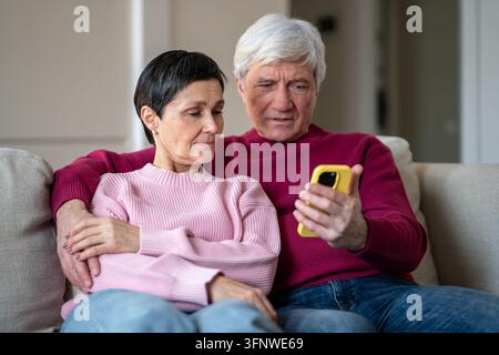 Positives, entspanntes Seniorenehepaar umarmt sich auf der Couch und schaut sich Medien auf dem Smartphone online an. Stockfoto