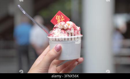Nahaufnahme einer Hand, die eine Tasse mit rosa gefrorenem Dessert hält, das mit Sahne und einer roten festlichen Dekoration in Singapur gekrönt ist Stockfoto