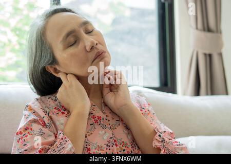 Nahaufnahme einer reifen asiatischen Frau, die ihren Hals hält und ihre Kehle berührt, Lymphknoten mit geschlossenen Augen in Unbehagen. Stockfoto