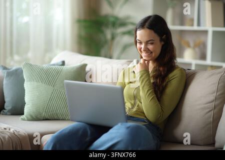 Nachdenkliche glückliche junge Frau in Grün, die einen Laptop benutzt und auf einer Couch zu Hause sitzt Stockfoto