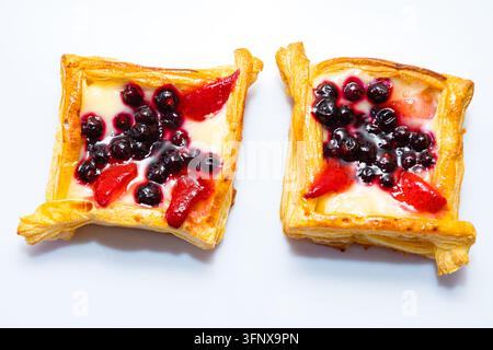 Studioaufnahme auf weißem Hintergrund von zwei quadratischen Blätterteigtörtchen, die mit Vanillesauce, Beeren und Erdbeerscheiben belegt sind. Knusprige gebackene Ränder umgeben glasiertes Stockfoto