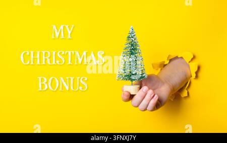 Eine Hand taucht durch zerrissenes gelbes Papier auf und hält einen kleinen Weihnachtsbaum. Studiofotografie mit lebendigen Farben. Geeignet für Feiertagsaktionen, fest Stockfoto