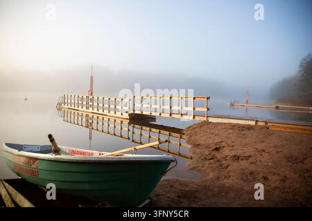 Ein Rettungsruderboot und eine Rettungsboje markieren einen offiziellen Badeplatz an einem nebligen finnischen Seeufer bei Sonnenaufgang, in der Nähe eines ruhigen Holzstegs. Stockfoto