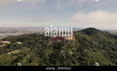 Luftaufnahme des Nationalpalastes von Pena, umgeben von üppigen grünen Wäldern und Hügeln mit der entfernten Küstenlinie, die unter einem weichen Himmel in Sintra sichtbar ist Stockfoto