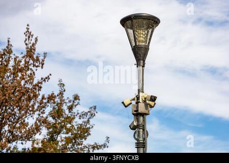 Ein metallener Straßenlaternenpfahl, der mit mehreren Überwachungskameras ausgestattet und mit Spinnweben bedeckt ist, steht vor einem teilweise bewölkten Taghimmel. Ein Stockfoto