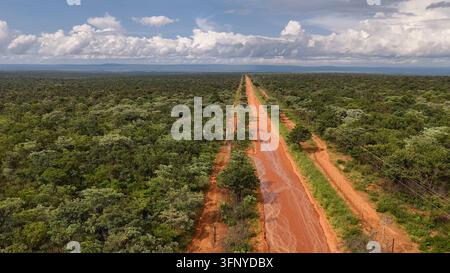 Luftaufnahme einer langen, abgelegenen roten Schotterstraße, die sich nach starken Regenfällen durch die abgelegene afrikanische Wildnis schlängelt, mit schlammigen Pfützen und üppiger Vegetation darin Stockfoto