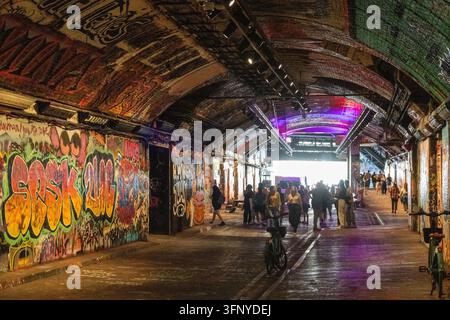 Ein geschäftiger gewölbter Ziegelgang in der Leake Street in London, bedeckt mit farbenfrohen Graffiti und Wandmalereien, mit Fußgängern, einem Fahrrad und lila und blauem Liguster Stockfoto