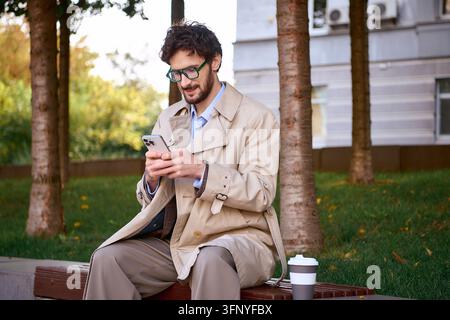 Ein junger, gutaussehender Geschäftsmann in schicker Kleidung überprüft sein Telefon im Freien in der Stadt. Stockfoto