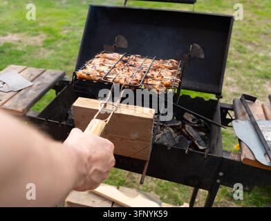 Ein konzentrierter Grillmeister hält einen Fleischspieß über einem Grill in einer Nahaufnahme. Geschickte Hände pflegen den Grill mit selbstsicherer Präzision und zubereiten Kebab im Freien auf einem sonnigen Holztisch. Stockfoto