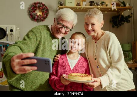 Großeltern mit Enkelin nehmen Selfie mit Kuchen auf dem Smartphone Stockfoto