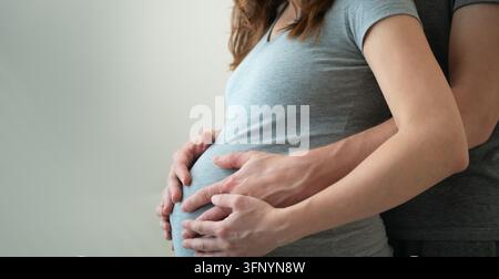 Schwangere Frau mit Partner, der seine Hände über ihren Bauch legt, in einem gemeinsamen intimen Moment. Erwartende Mutter und Ehemann umarmen sich von der Seite im Studio mit neutralem Hintergrund. Horizontales Banner Stockfoto