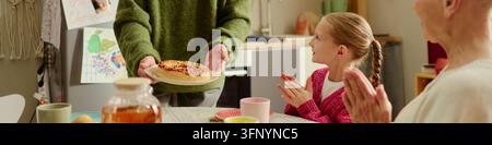 Kopfzeile von Mann, der frischen Kuchen an ein glückliches Mädchen mit einer Seniorin am Tisch serviert Stockfoto